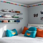 Bedroom corner with monorail-inspired silver wall shelves displaying repainted toy boats and buses, gondola-shaped throw pillows in blue and orange on a daybed, and a slim washi-tape monorail track tracing the ceiling edge in soft natural light.